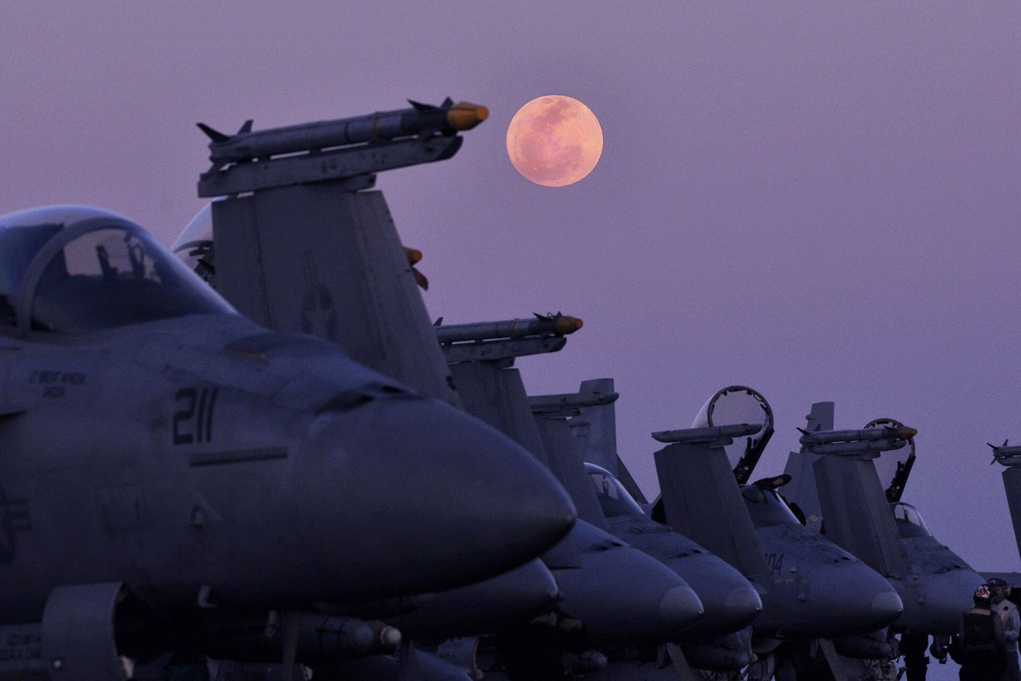 A row of armed fighter aircraft parked on a carrier flight deck at dusk, a full moon rising behind them.