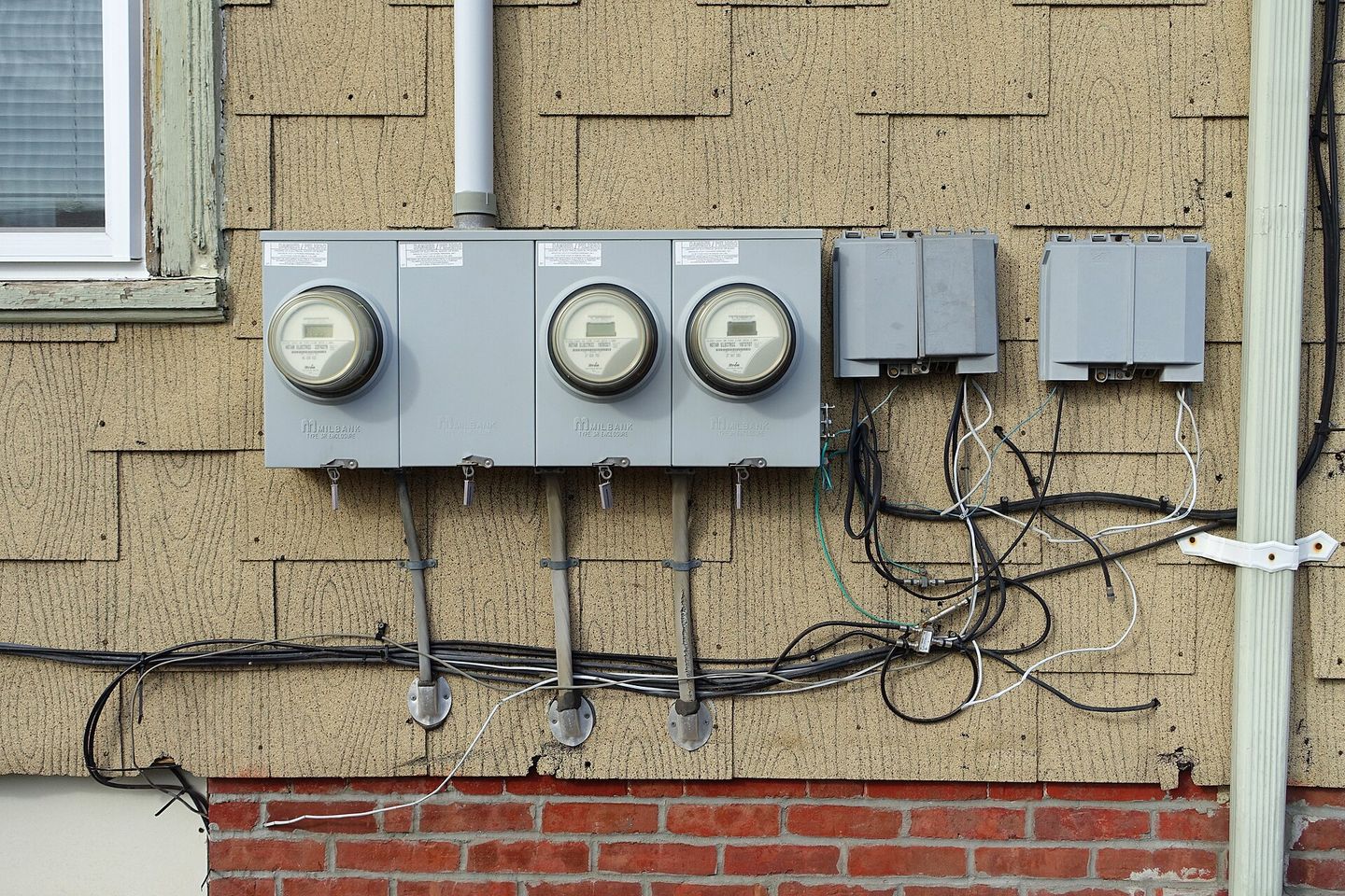 A bank of four domestic electricity meters on the shingled wall of one building, conduit and cable running away from them.