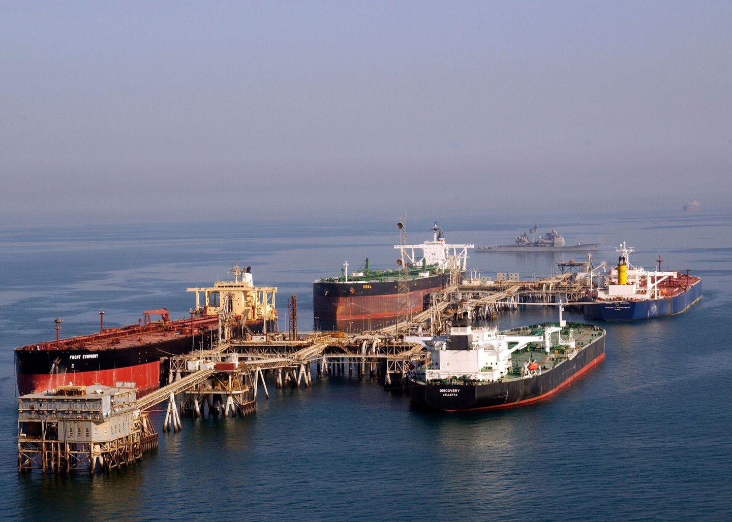 Trois pétroliers amarrés à un terminal de chargement au large, dans le Golfe, un bâtiment de guerre à l'horizon.
